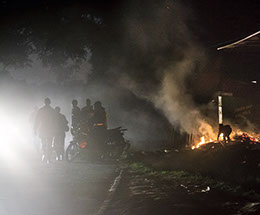 Child fuels bonfire, while silhouetted men stand around a motorcycle. Smoke, headlights